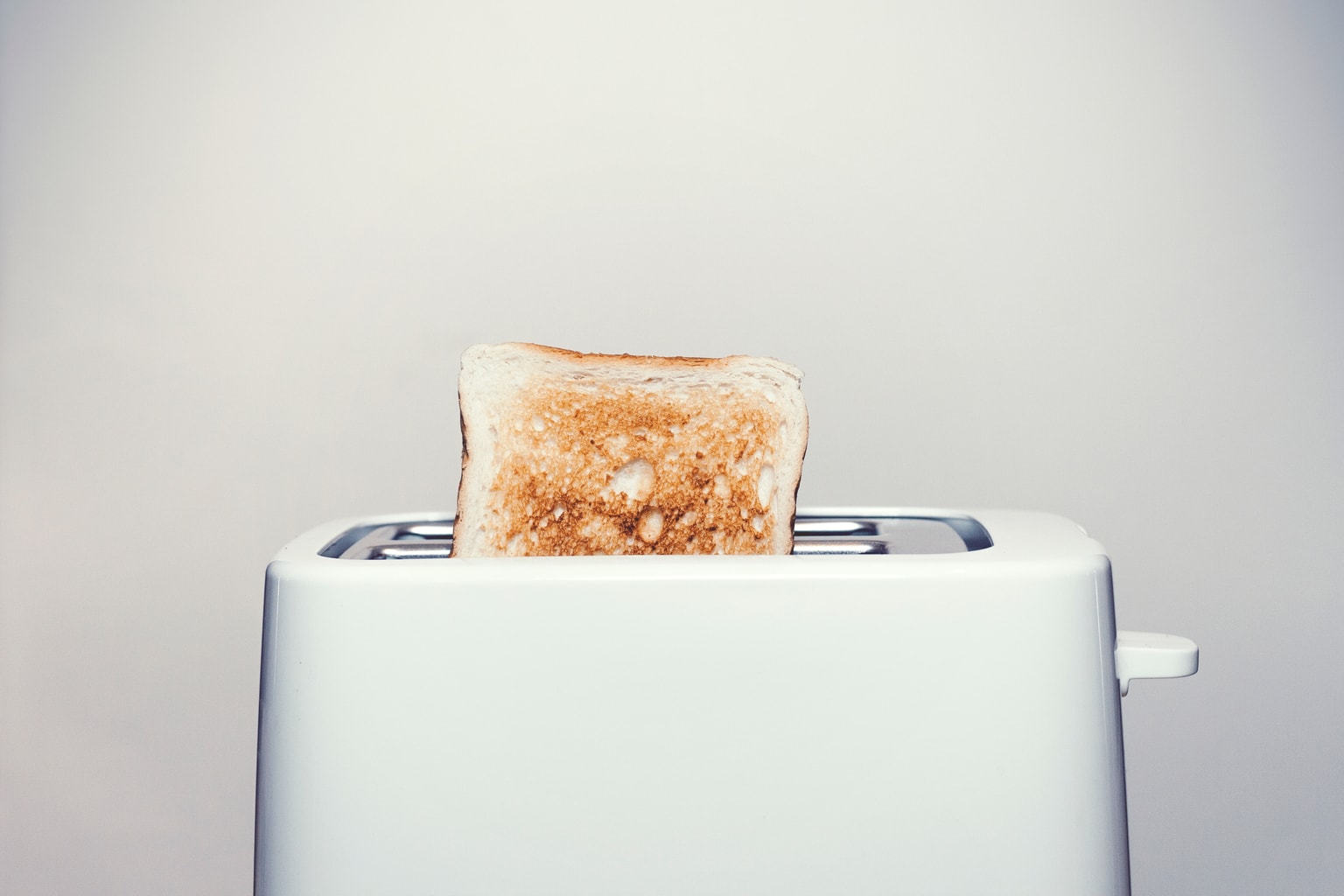A piece of toast popping out of a white toaster against a gray background.