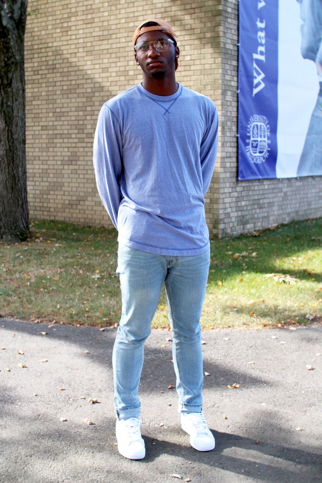 University of Bridgeport student Brandon wears a simple blue long-sleeve t-shirt, skinny-fitted light-wash denim jeans, bright white Adidas sneakers, and a rustic orange baseball cap.