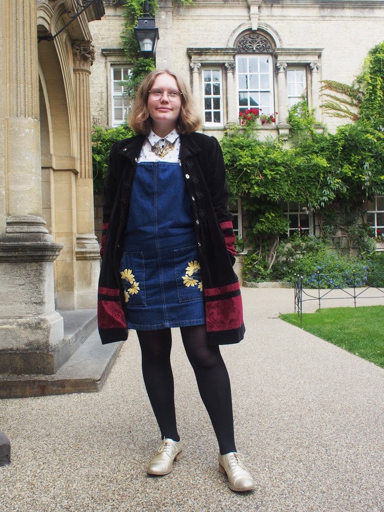 Hannah wears a denim jumper dress with embroidered pockets, a white collared button-up shirt, and a vintage long black coat with maroon detailing.