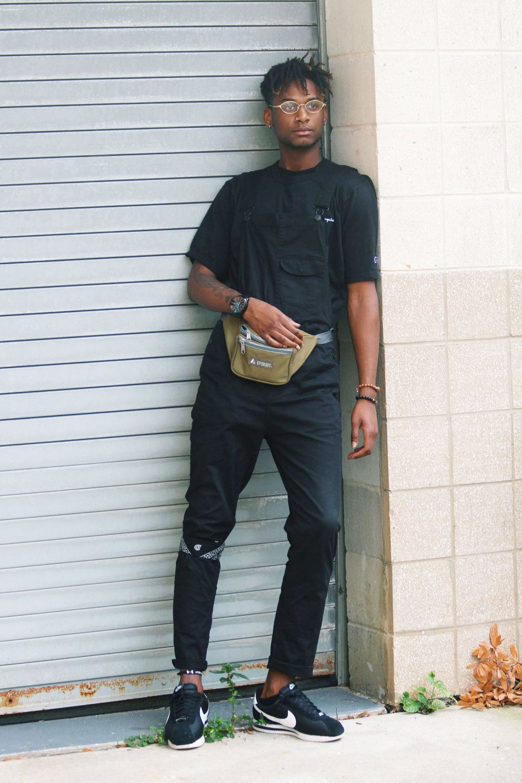 USF Fashion: Mens college fashion at University of South Florida - student Karon wears an all black outfit including overalls, a bandana, a black tee shirt, a fanny pack, glasses, and Nike sneakers