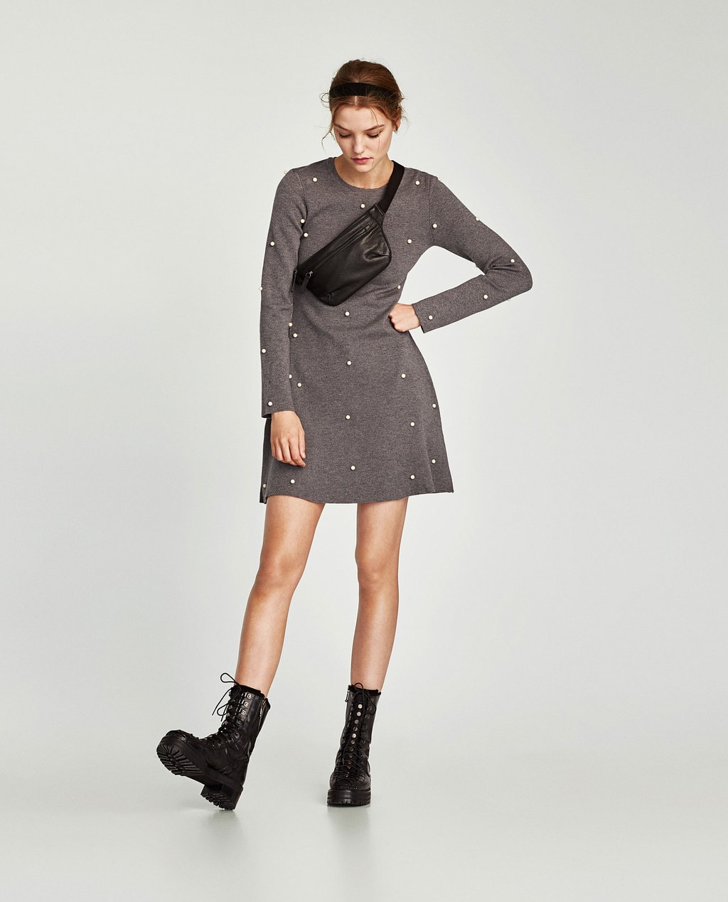 Photo of model standing against light gray background wearing medium-dark gray heathered long-sleeved skater dress with white pearl embellishments, black combat boots, a black pebbled leather fanny pack across one shoulder, and a black headband