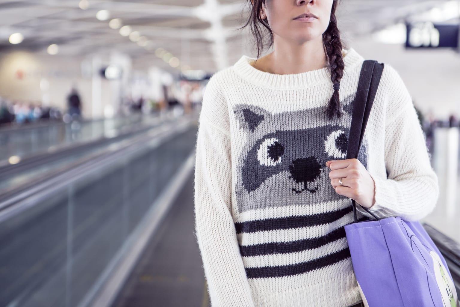 Woman at the airport on a moving sidewalk wearing a raccoon sweater and carrying a purple tote bag