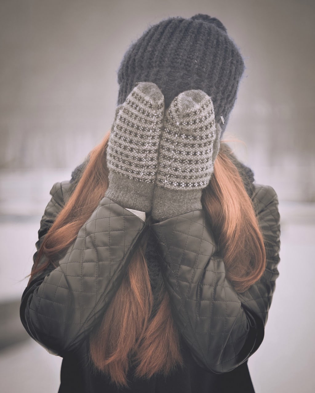 Woman wearing mittens and a quilted leather jacket, covering her face in the winter snow
