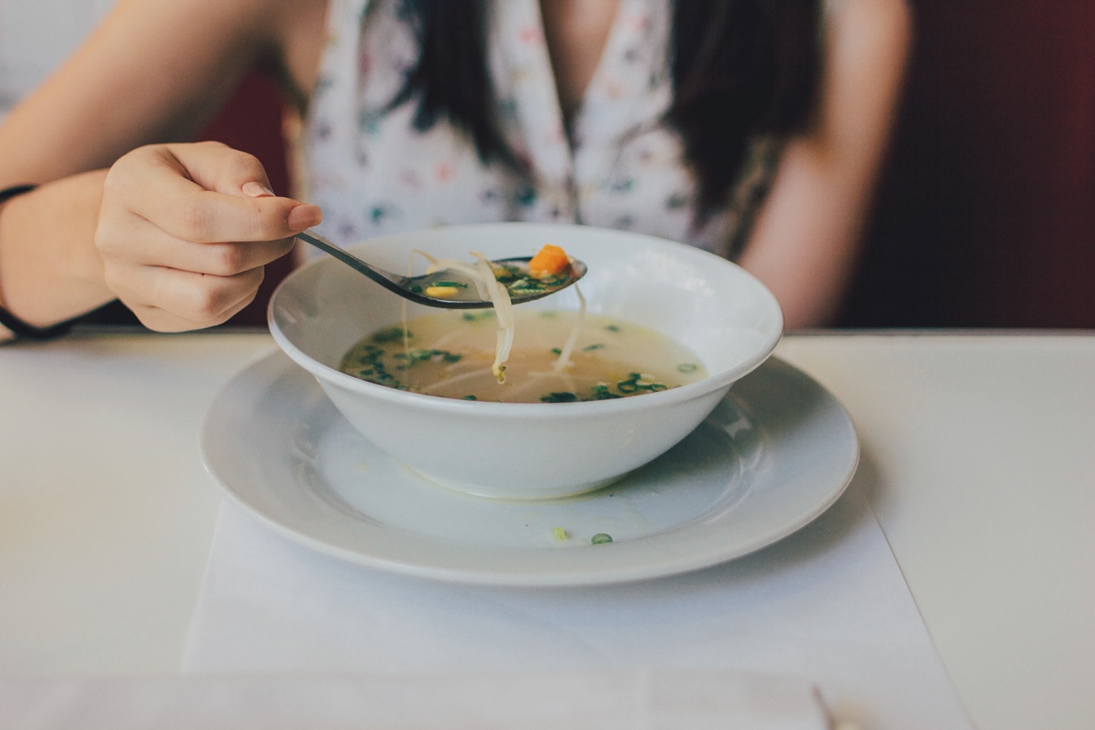 A seated woman, her face out of view, holds a spoonful of soup above a bowl.