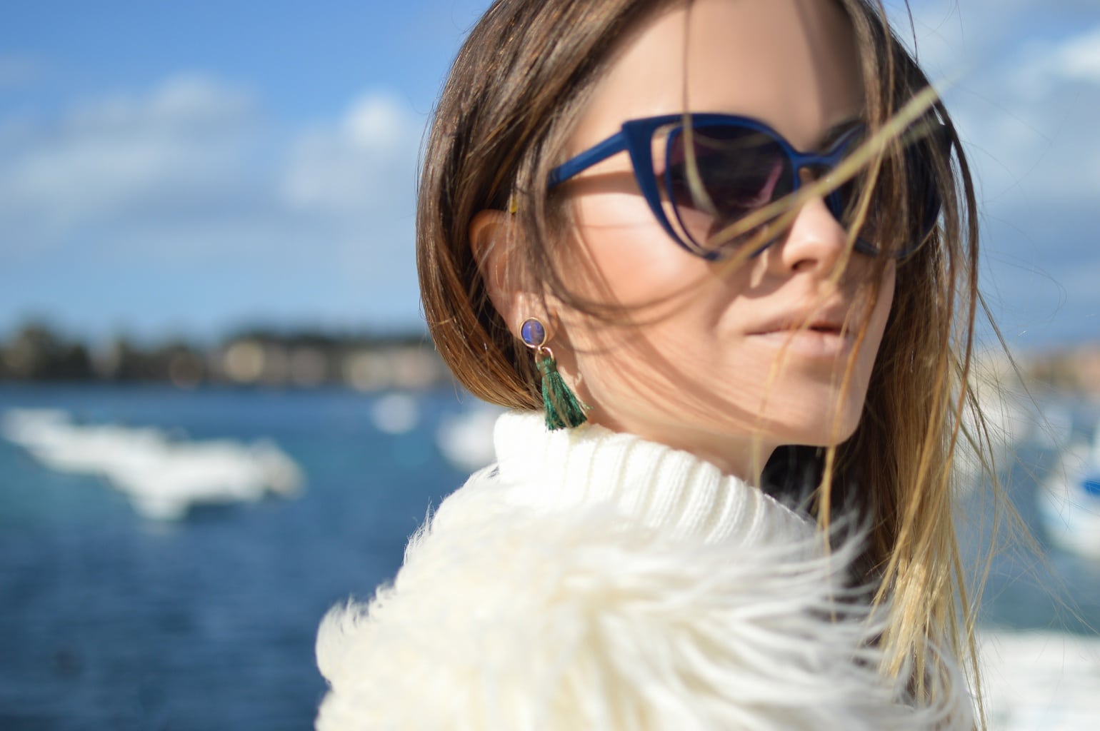 Single woman standing in front of the ocean, wearing sunglasses and a fur jacket