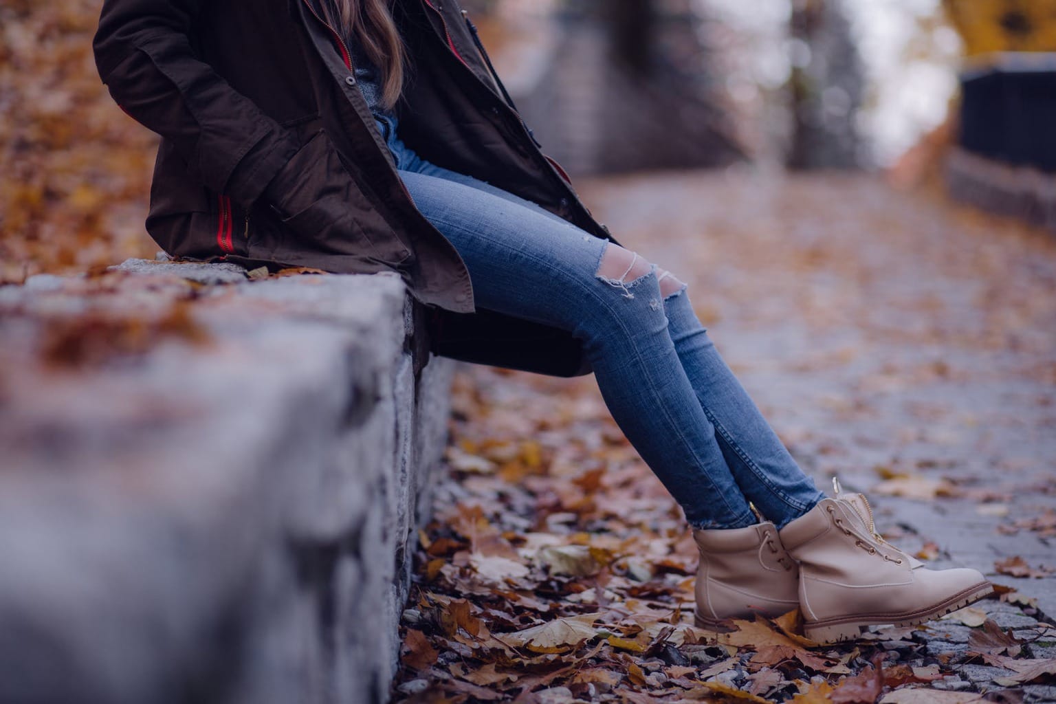 Woman sitting on a ledge in fall wearing blue jeans, boots, and a black jacket