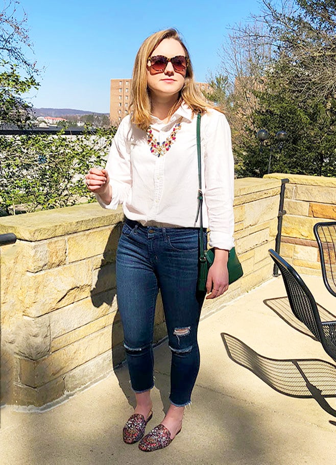 This West Virginia University student wears a basic white linen button-down shirt tucked into a pair of basic dark skinny jeans with distressed knees. She sports them with flat jeweled mules from Target and a green Kate Spade over-the-shoulder purse. She finishes off her look with a statement gold necklace full of red, baby blue, and navy blue jewels.