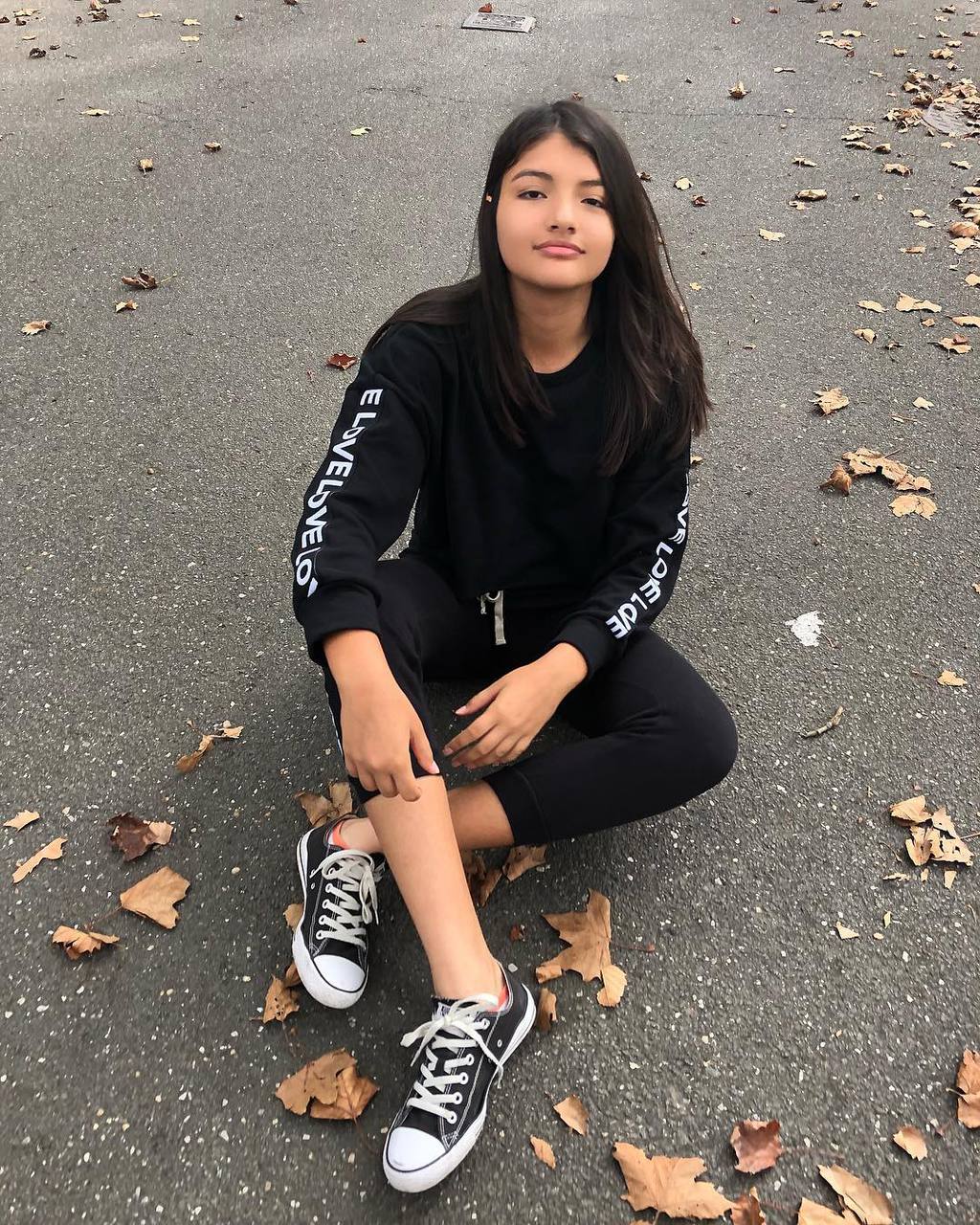 Nathaly wears a black matching sweatsuit with white words on the sleeves and matching black low-top Converse sneakers.