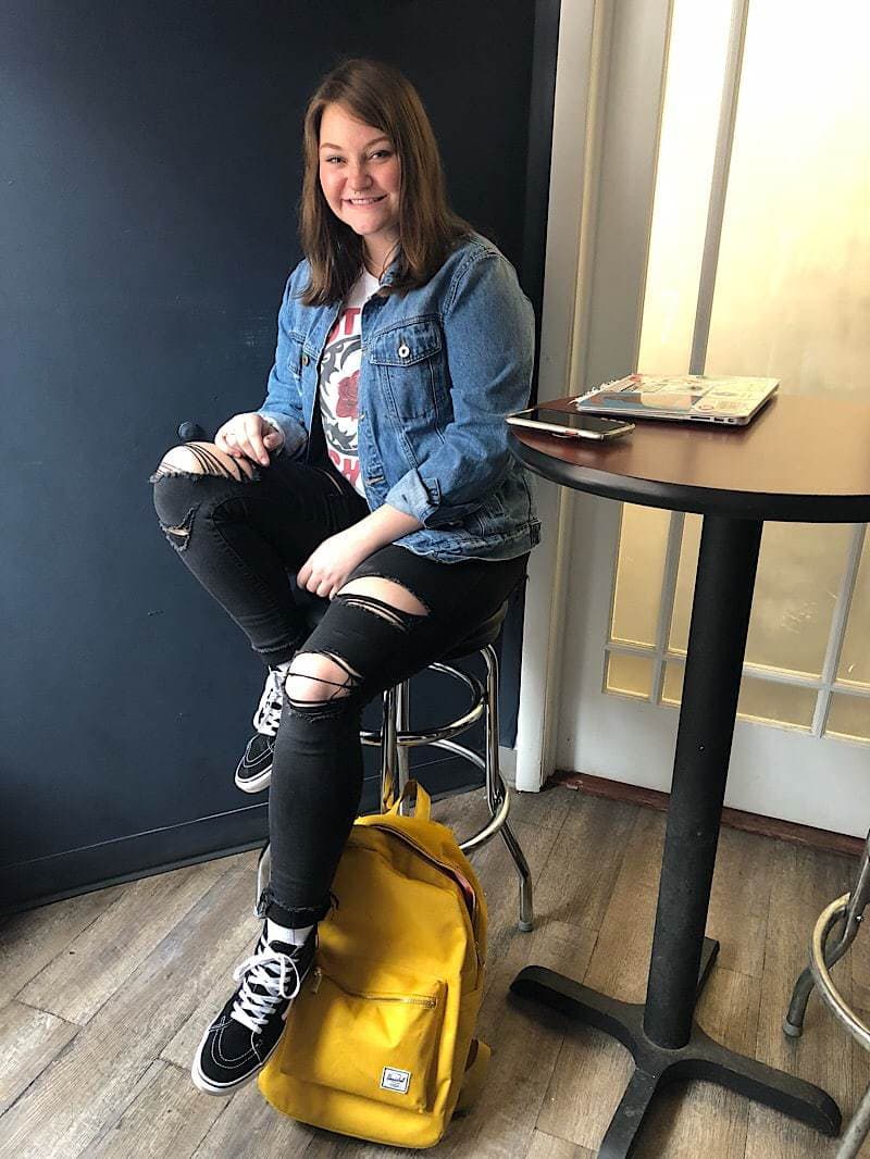 Tiana, a WVU student, wears a white graphic tee, ripped black jeans, Vans sneakers, and a medium-wash denim jacket.