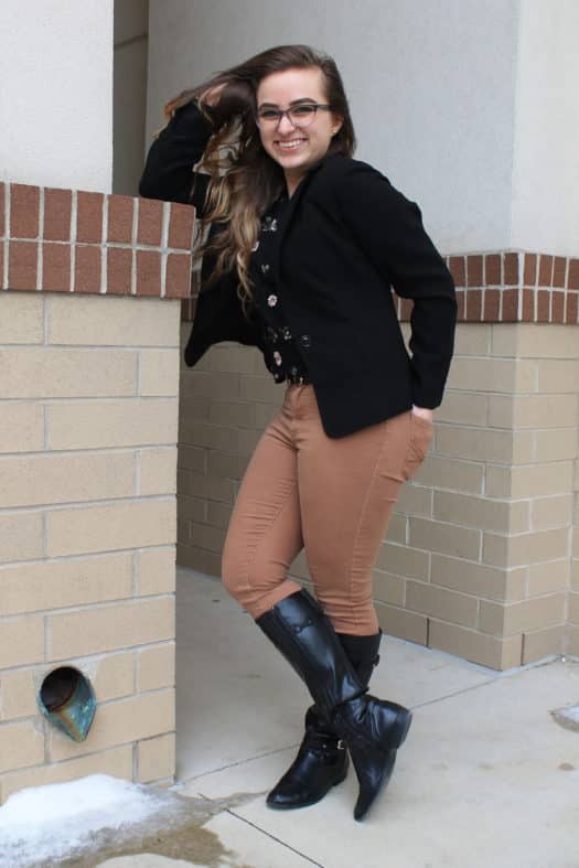 Leslie wears a black floral print shirt with a black blazer and camel colored jeans tucked into black riding boots.