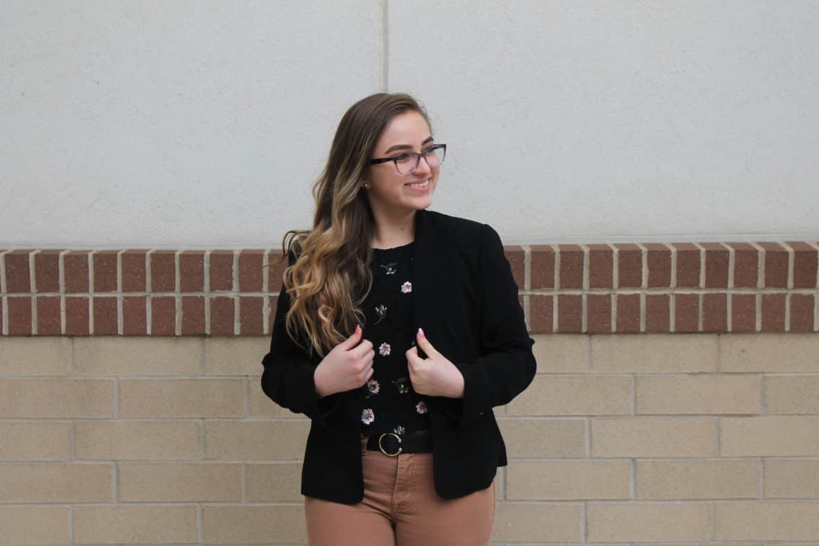 Leslie wears a fitted black blazer with a black floral print shirt tucked into her camel jeans.