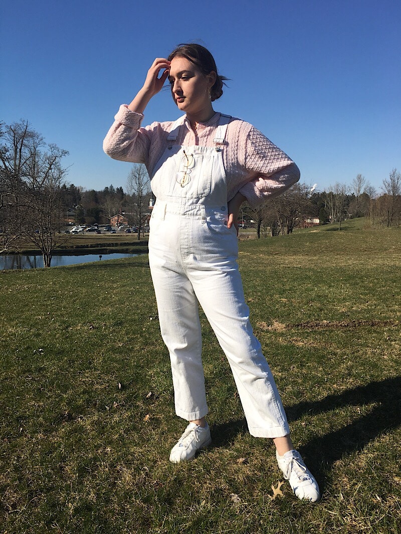 Abby wears cropped white denim overalls with a heavy cable knit baby pink sweater-shirt and white Keds.