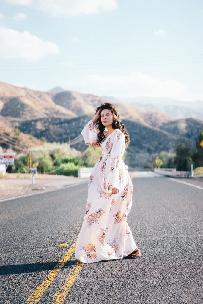 Woman in a long, floral-print dress standing on a road against a backdrop of mountains.