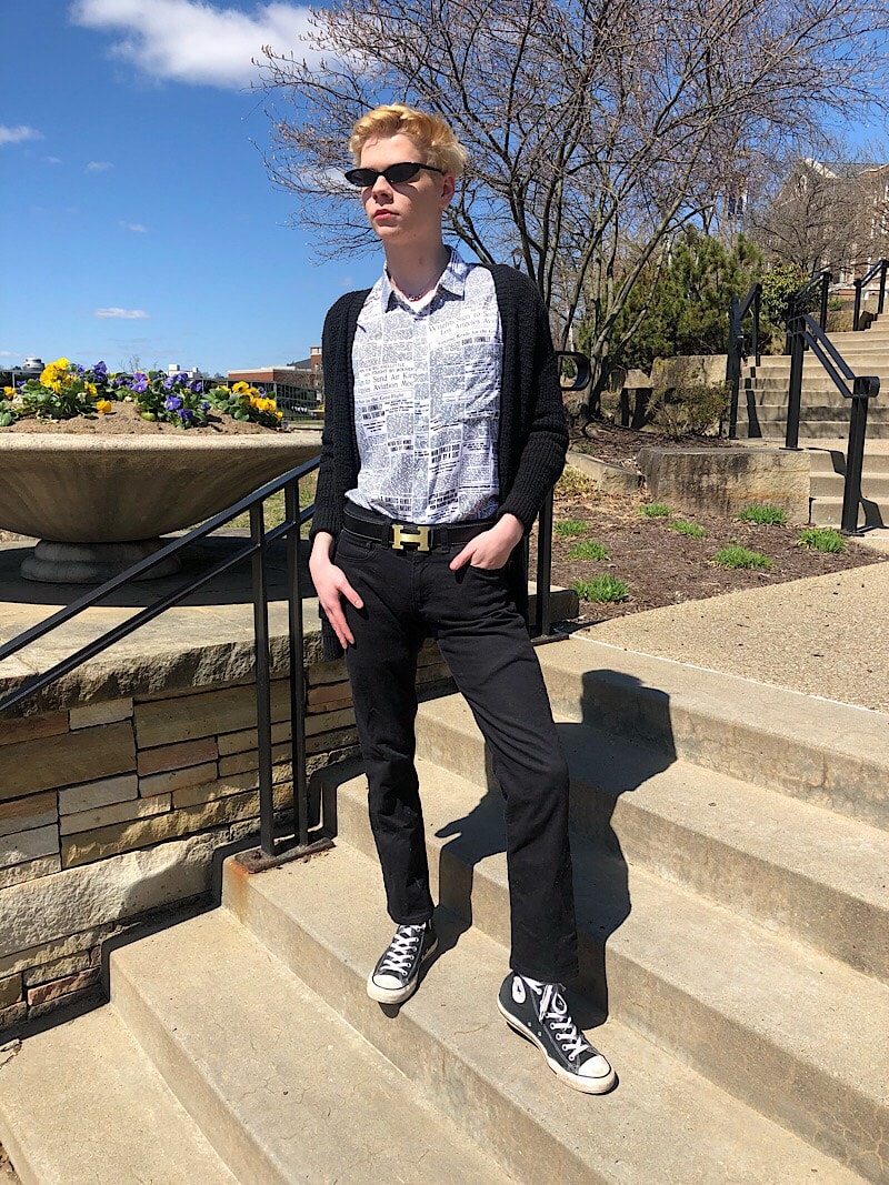 Caleb wears skinny black plastic framed sunglasses with his newspaper print button-up shirt, black sweater, black denim, classic Converse sneakers, and Hermes belt.