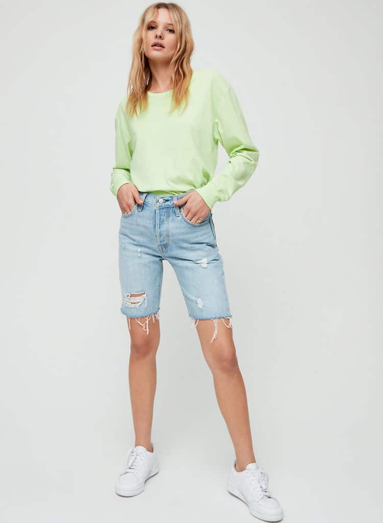 Woman with her hands partially in her pockets wearing a light neon green sweatshirt tucked into a pair of light-wash, distressed denim Bermuda shorts, white sneakers, and delicate gold rings on her little finger