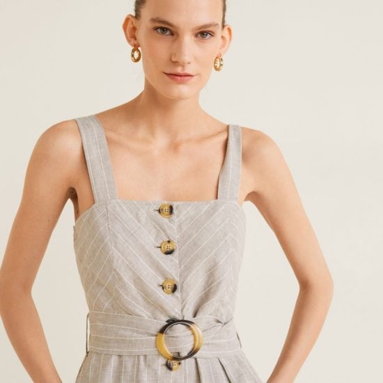Close-up image of woman standing against a beige background wearing small, thick gold hoop earrings and a sleeveless button-front belted jumpsuit in beige with thin white pinstripes.