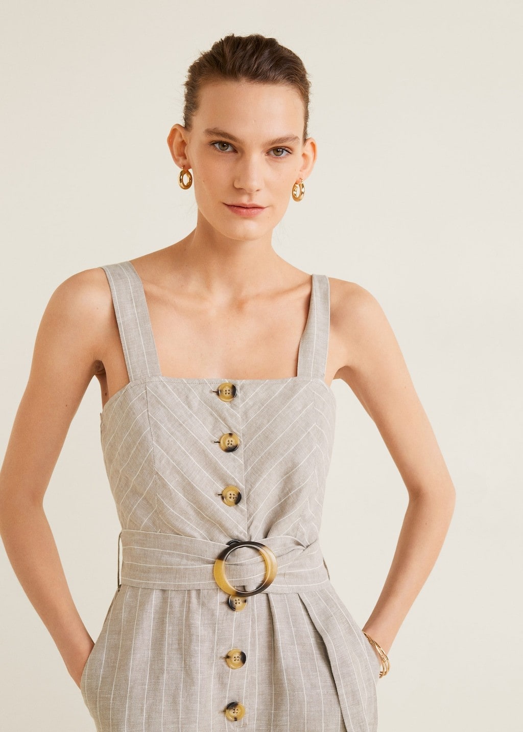 Close-up image of woman standing against a beige background wearing small, thick gold hoop earrings and a sleeveless button-front belted jumpsuit in beige with thin white pinstripes.