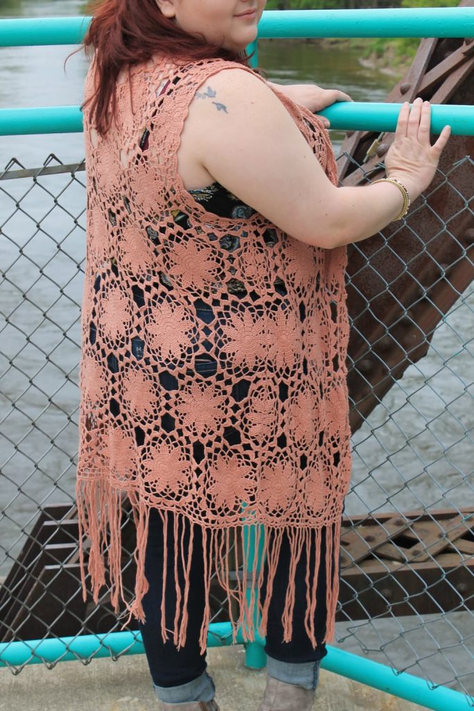 Brandi wears a long, coral, crochet duster vest with fringe at the bottom.