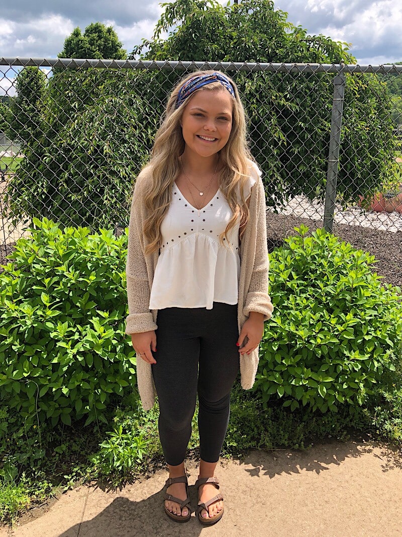 Kelsey wears a white ruffled top with rivet details, a long tan cardigan, black leggings, and brown birkenstock sandals.