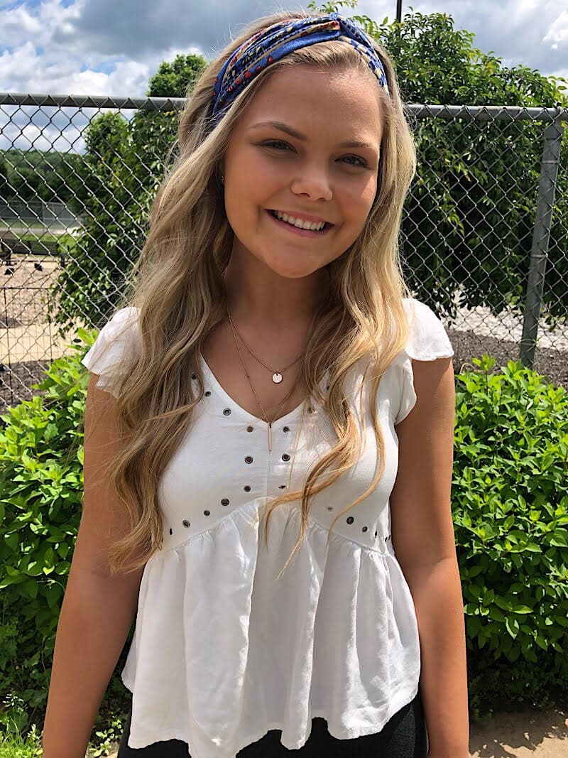 Kelsey wears a blue headband and a simple white babydoll top with flowy details and rivets.