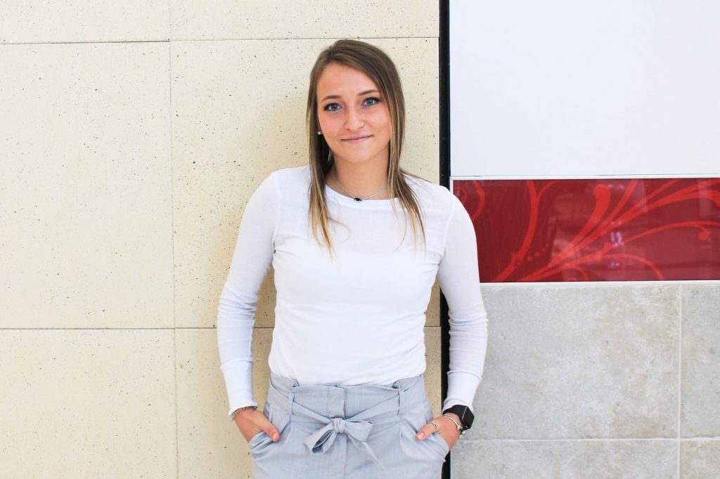 Darby wears a basic long sleeve white shirt tucked into her grey trousers.