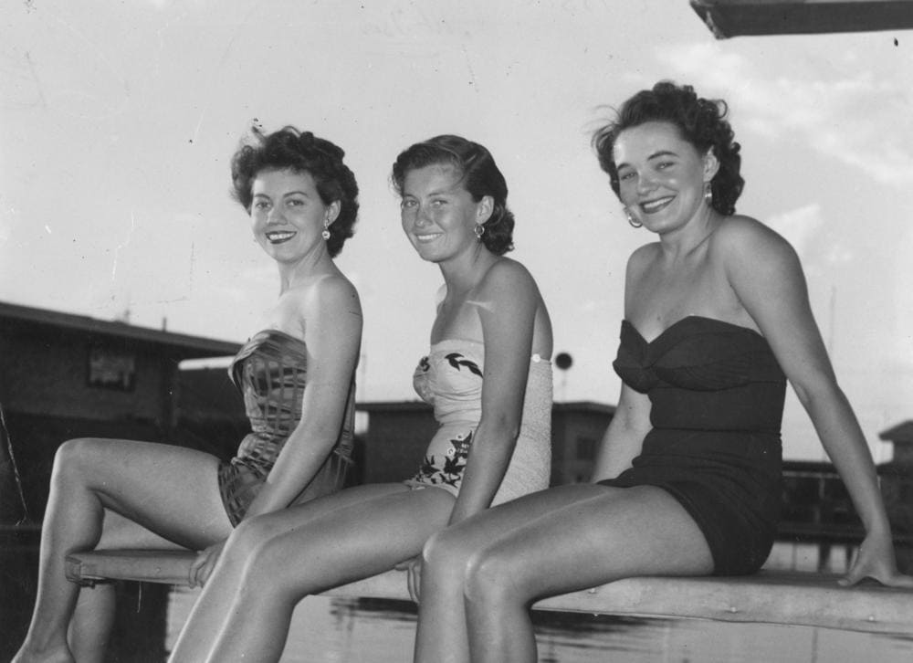 Three 1950s women sitting on bench wearing one-piece swimsuits