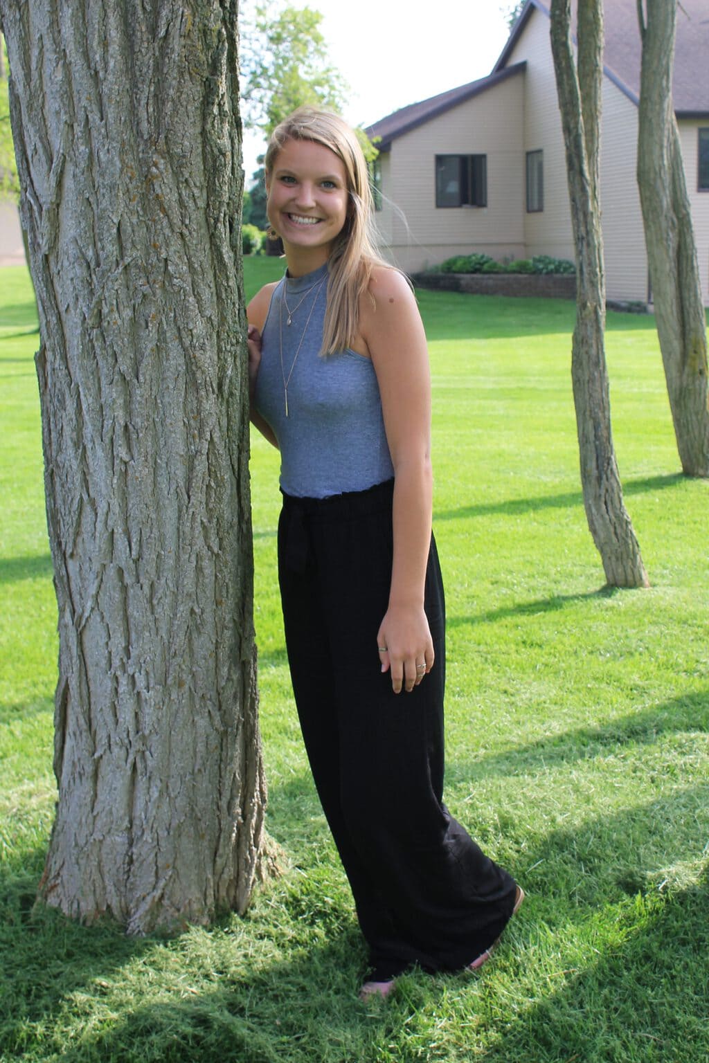 Emily wears a grey high-neck sleeveless tank top, flowy black pants, and layered gold necklaces.