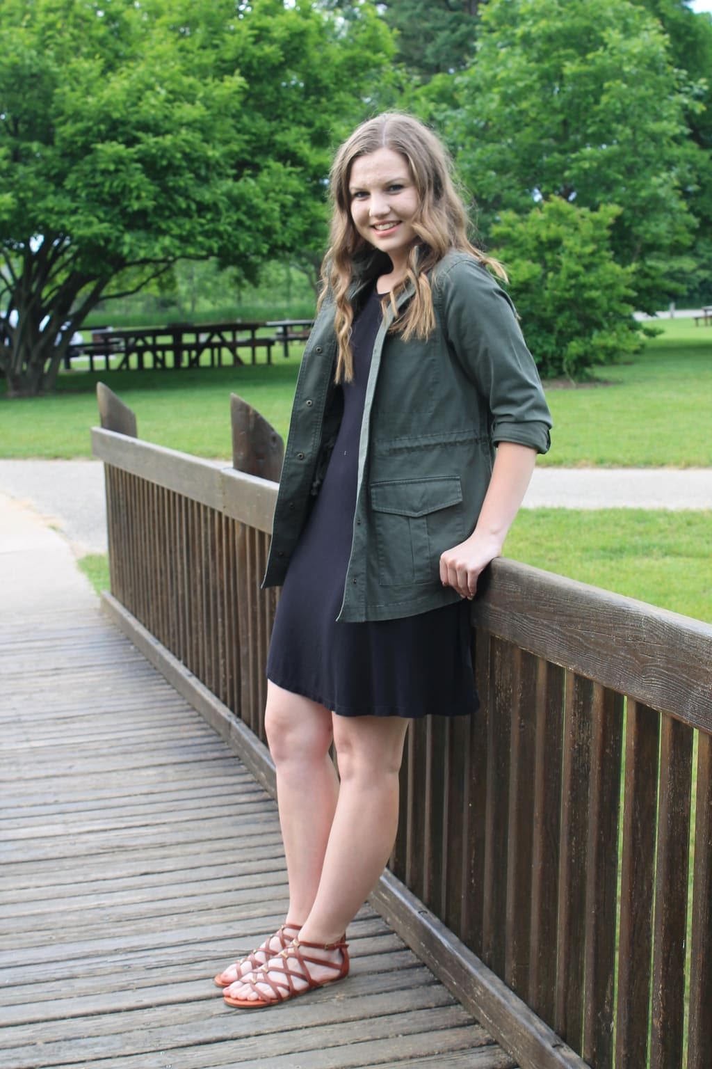 Rebecca wears an army green anorak jacket, a simple black shift dress, and brown criss-cross flat sandals.