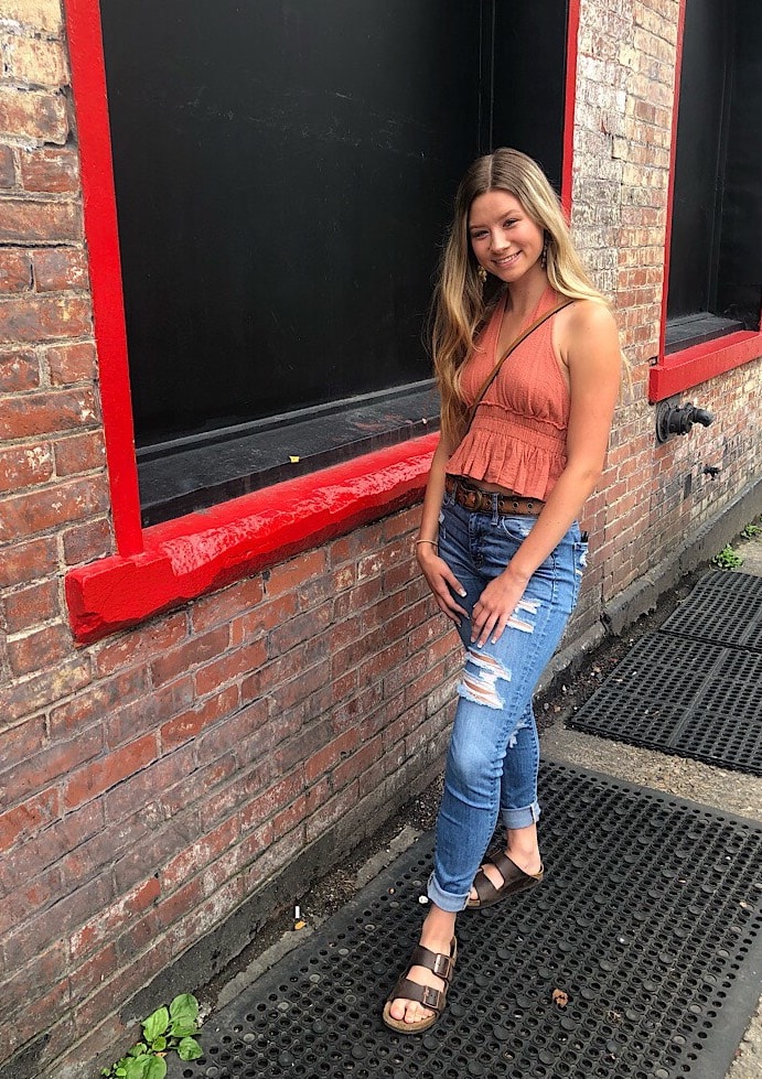 Logan wears a burnt orange halter top with a ruffle peplum, ripped and cuffed skinny jeans, and dark brown birkenstock sandals.