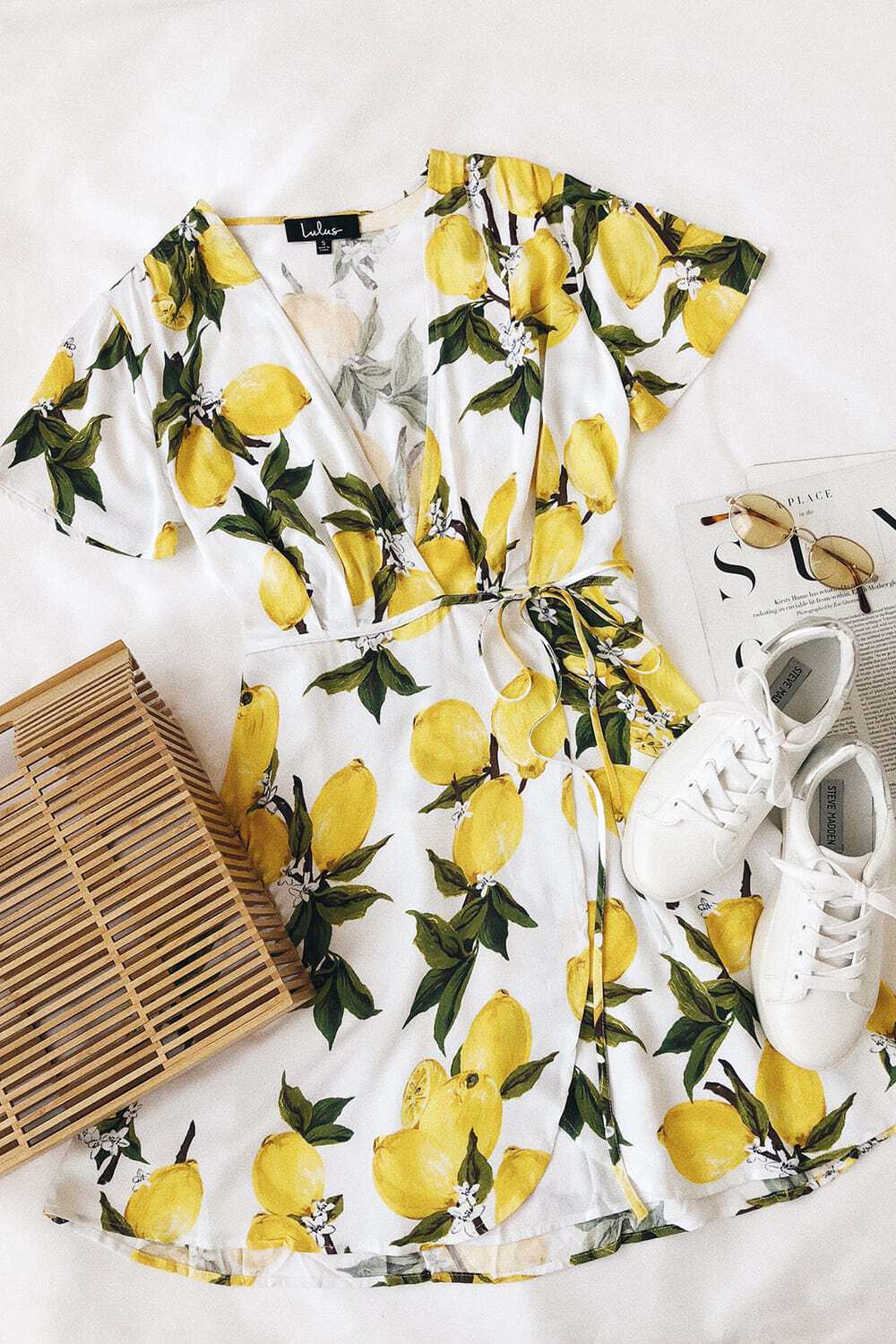 Flat lay featuring short-sleeved lemon print wrap dress, wooden bag, small oval sunglasses, and white sneakers, with part of an open magazine visible in the background