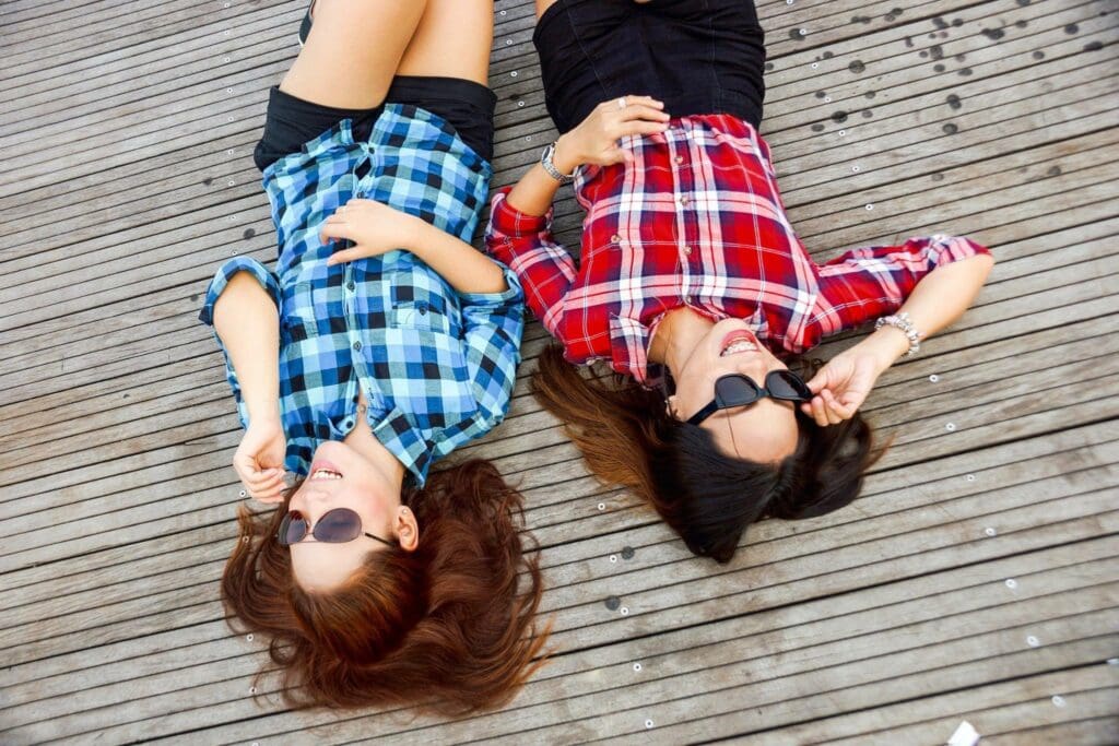 Two women in plaid shirts and sunglasses, laughing and laying on their backs on the ground.