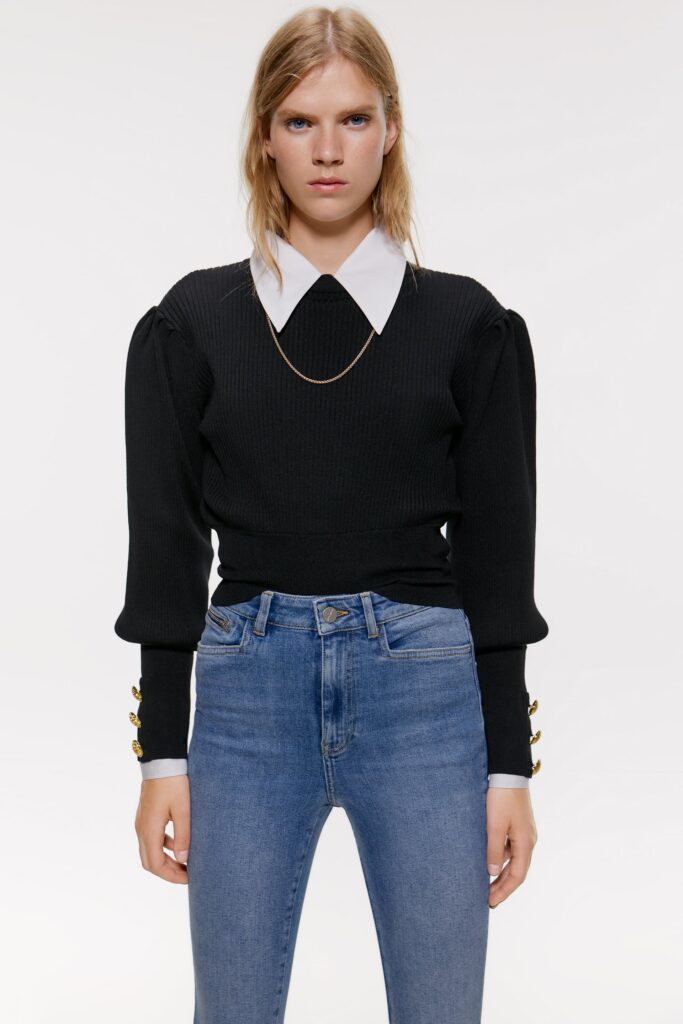 Young woman wearing a black puff-sleeve sweater with three gold buttons at each cuff over a white shirt with an oversized collar, as well as a skinny gold chain necklace and light-wash skinny jeans