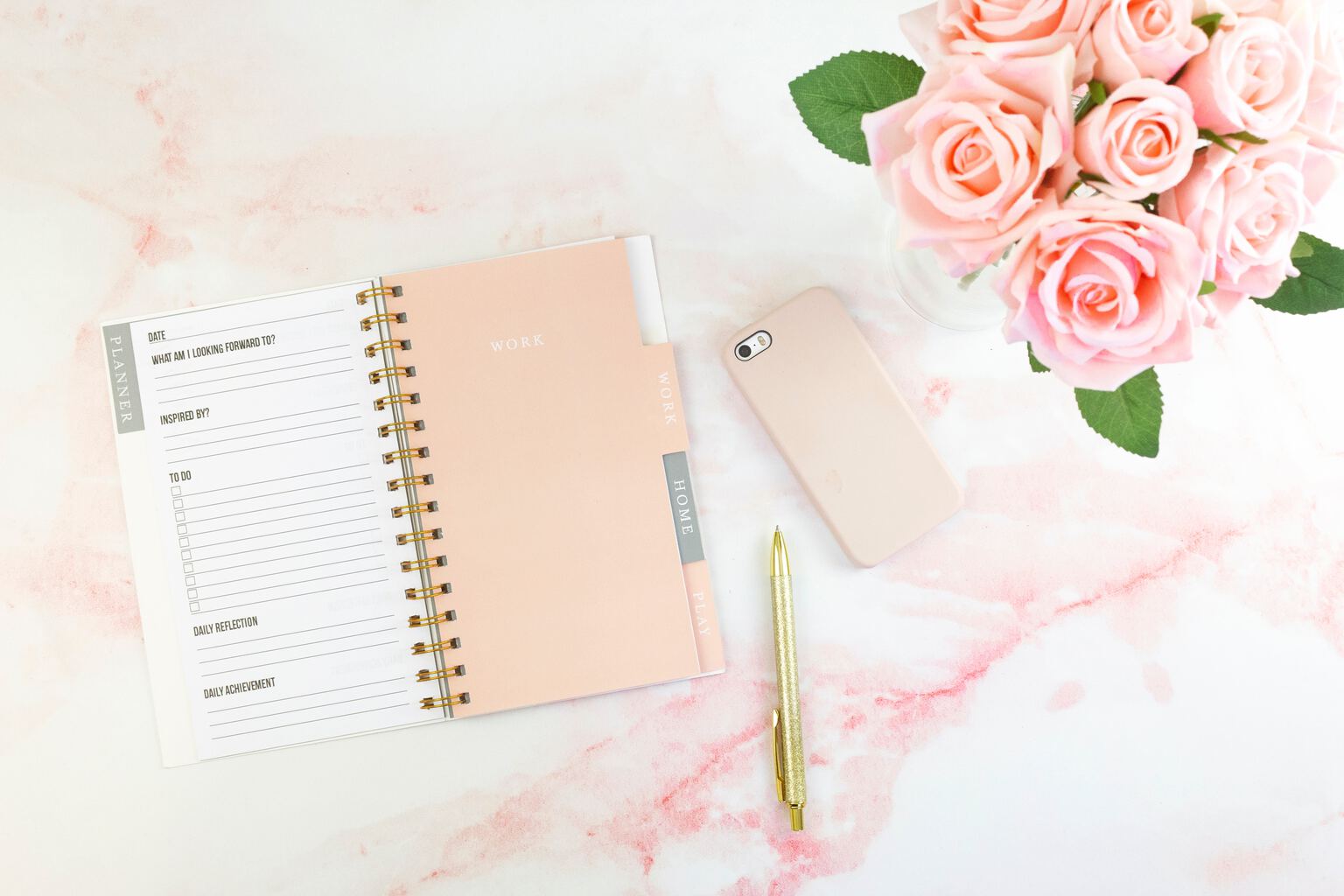 Flat lay on light pink marble surface featuring planner, gold-colored pen, iPhone in pale pink case, and pale pink roses