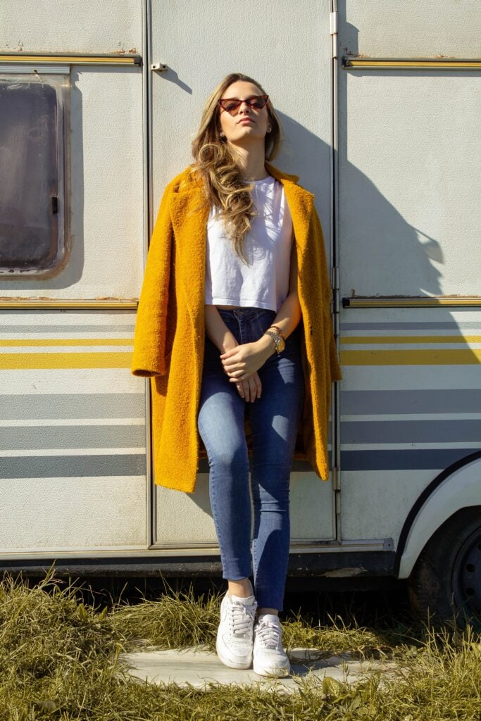 Woman standing outside in front of vehicle wearing yellow coat over white boxy-fit t-shirt and skinny jeans, as well as a watch and a bracelet, earrings, cat-eye sunglasses, and white sneakers