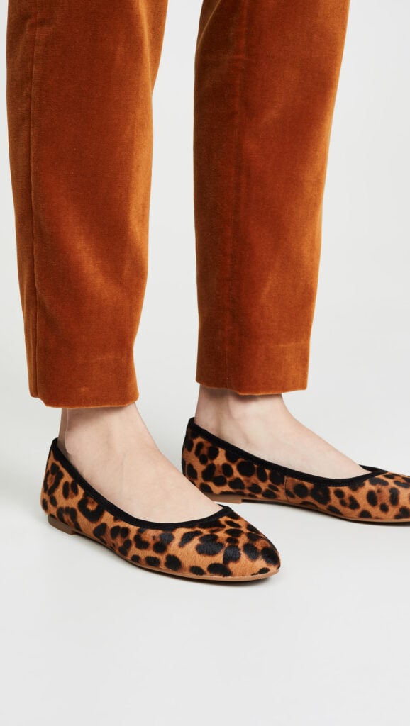 Shot of model's legs from the shins down; model is wearing dark, warm beige slim-fitting pants and calf-hair leopard-print ballet flats with black trim