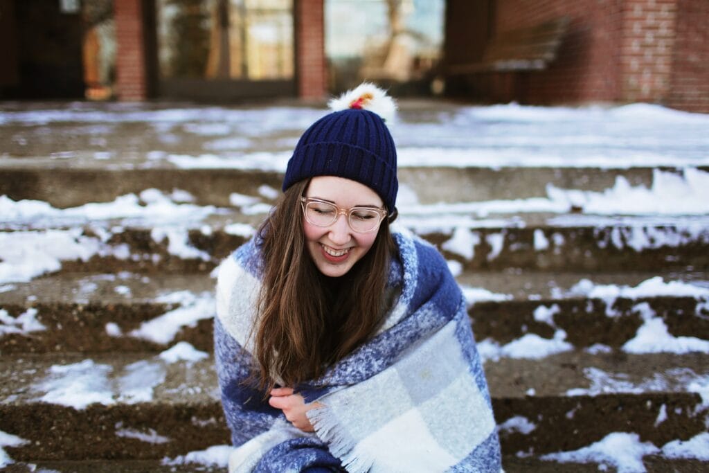 Girl in blanket and hat sitting on steps in the snow.