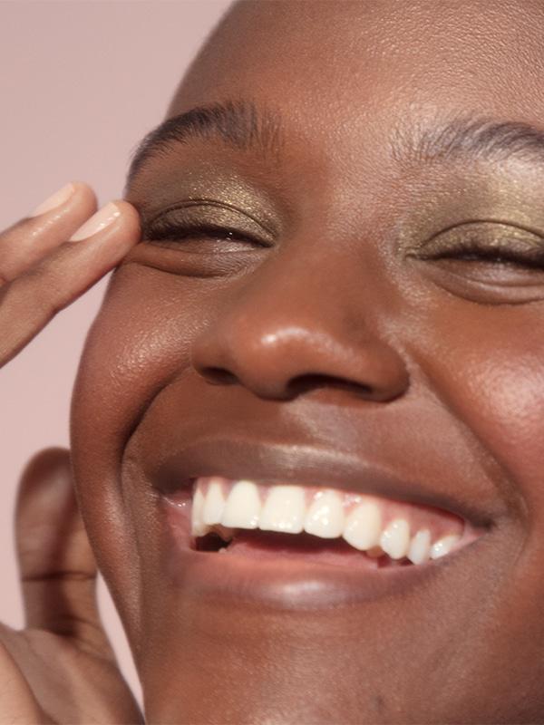 Close-up shot of woman smiling against pale pink background and wearing Glossier Lidstar in "Herb." She is gently touching the corner of her right eye with the finger of her right hand.