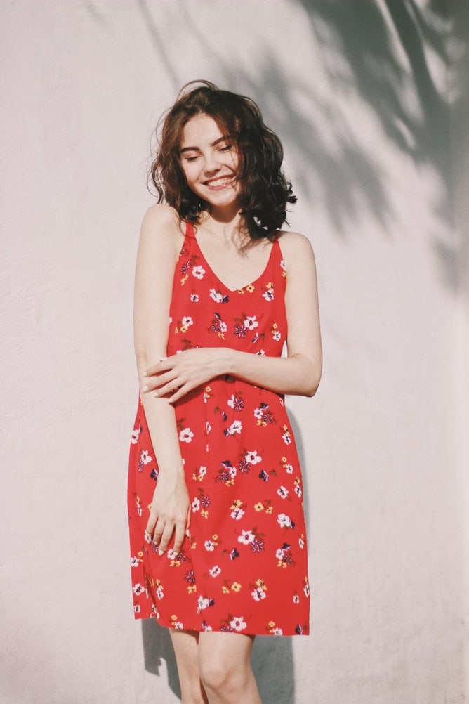 Smiling young woman wearing sleeveless red floral-print dress standing against white wall outside