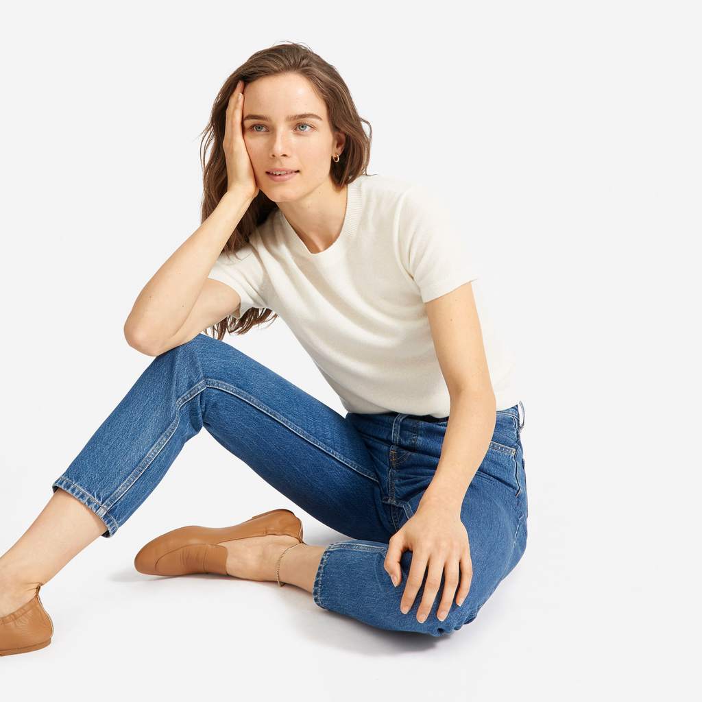 Woman sitting on the ground with her head in her hand wearing an ivory-colored short-sleeved sweater, cropped medium-wash jeans, and cognac-brown loafers