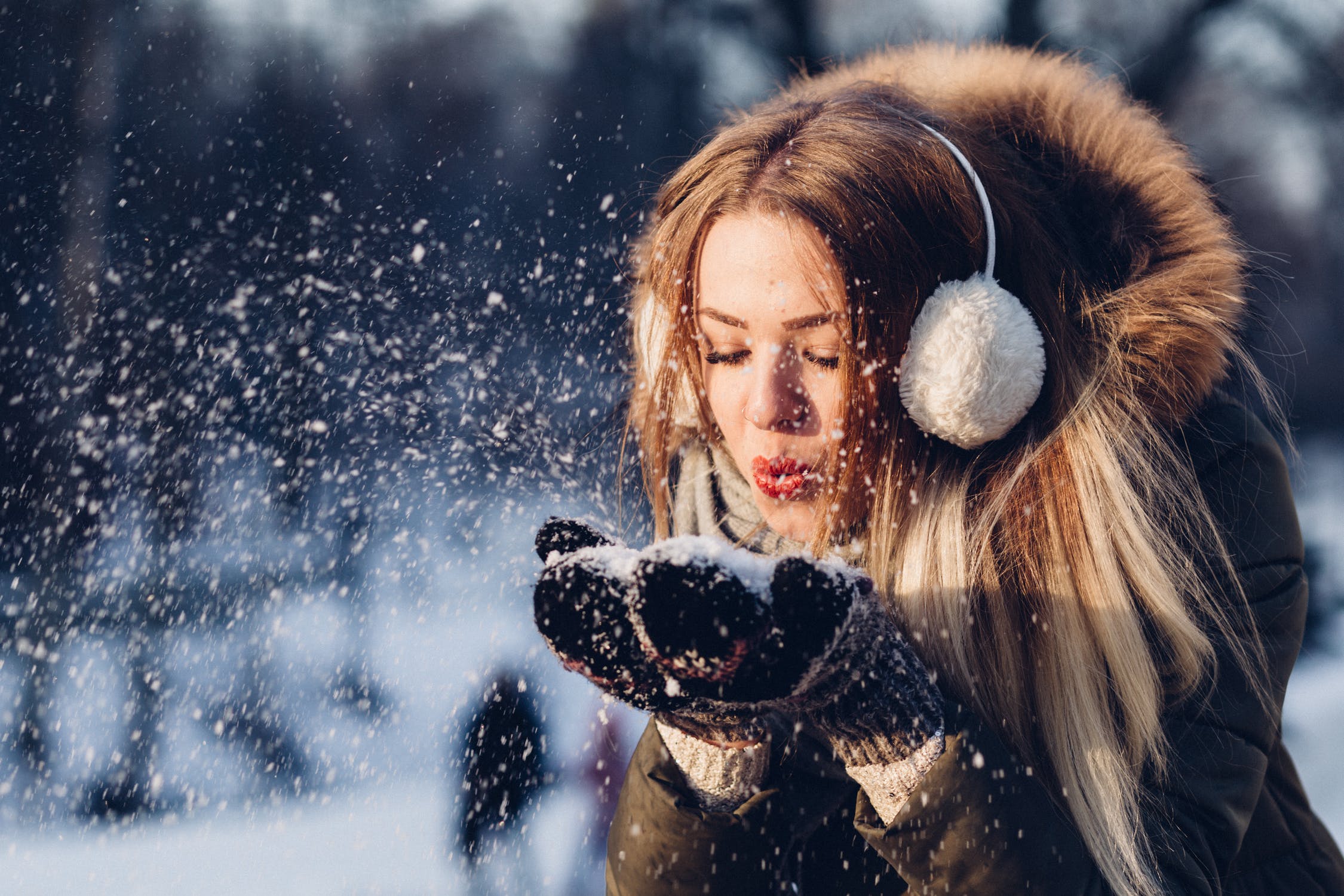 Woman blowing snow wearing earmuffs and a fur hood.