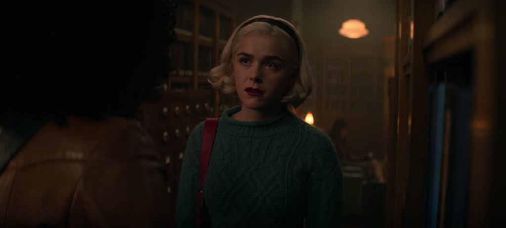 Screenshot from episode 1 of The Chilling Adventures of Sabrina Spellman, Season 4 - Sabrina in a green sweater and headband inside a library