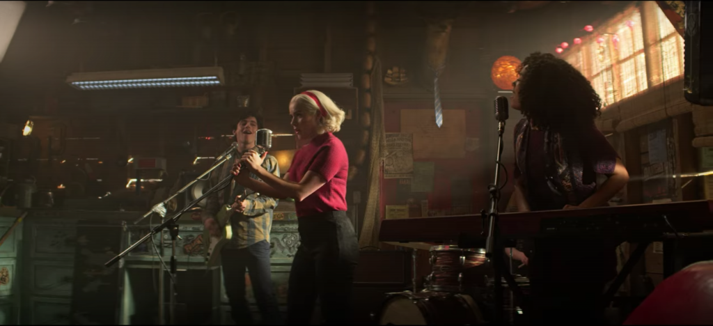 Screenshot from episode 5 of The Chilling Adventures of Sabrina Spellman, Season 4 - Sabrina performing in a garage in a red sweater and matching headband