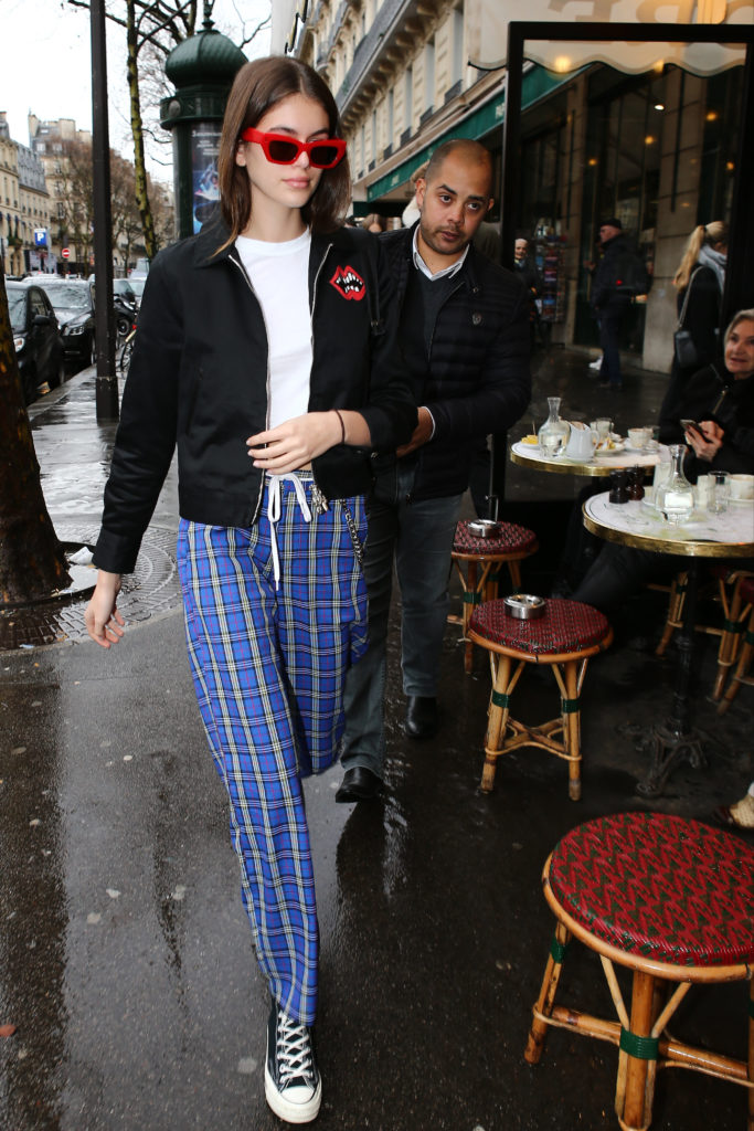 Kaia Gerber wearing blue plaid pants, black converse sneakers, a black zip jacket, and red sunglasses