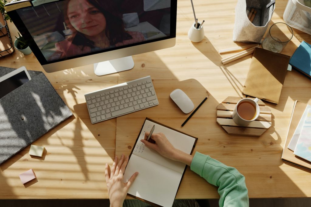 Picture of a woman writing in a notebook in front of a computer screen.