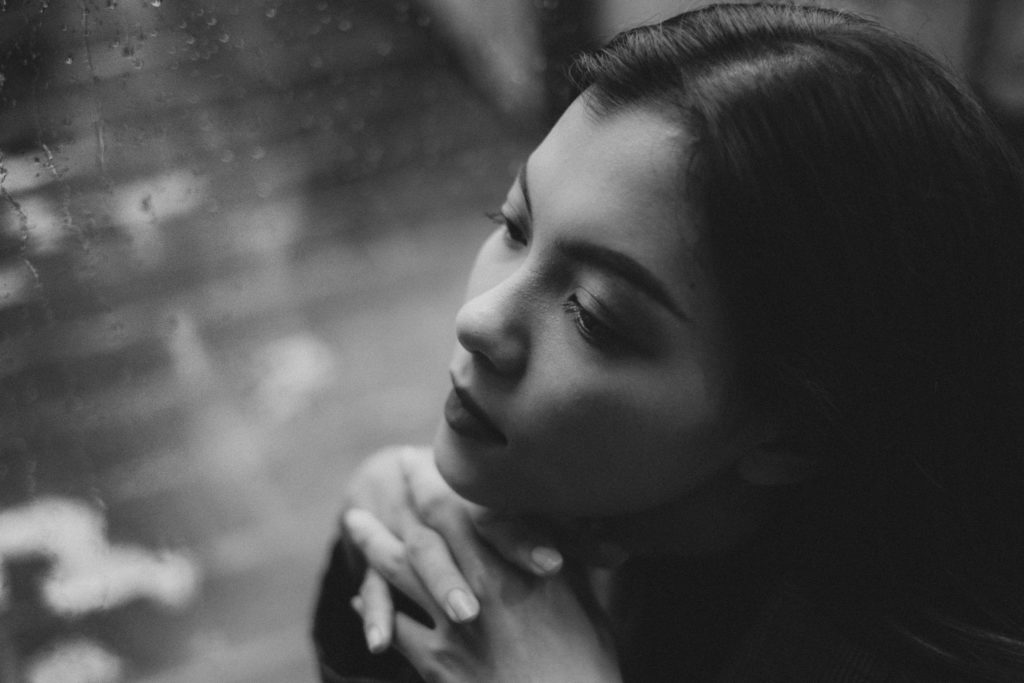 A black and white picture of a girl sitting by a rain covered window.