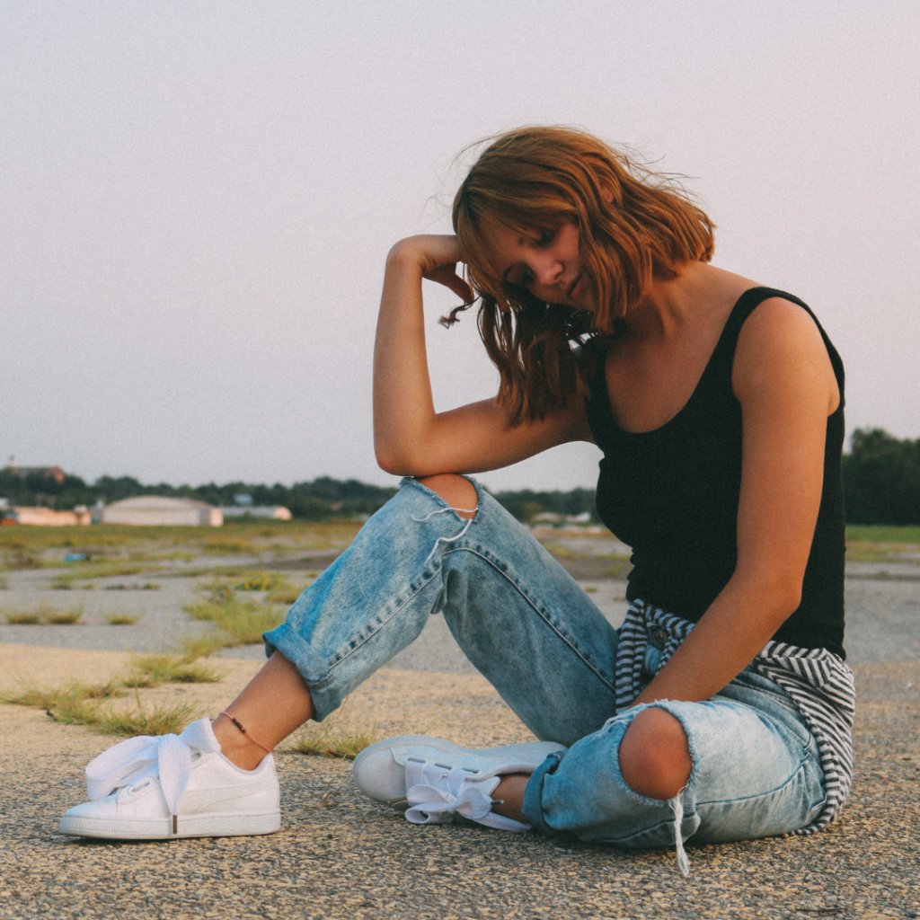 Woman wearing ripped jeans, white sneakers, and a white tank top while sitting on the ground in a parking lot