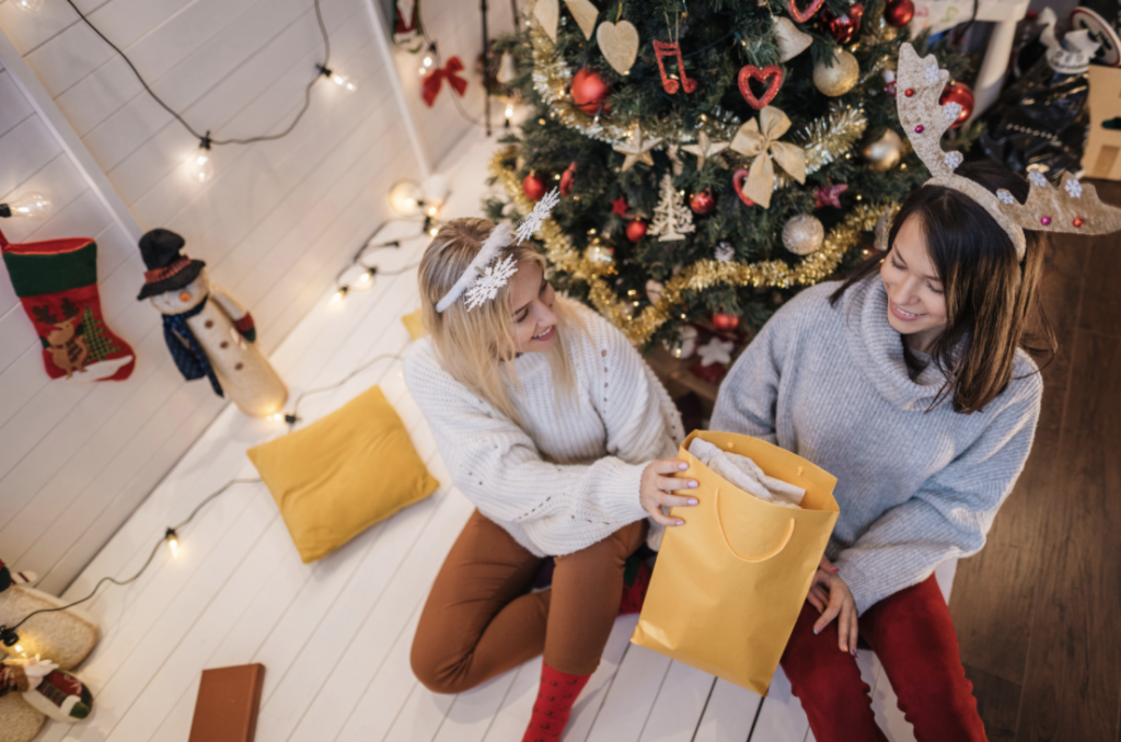 Two women giving each other gifts while sitting near a Christmas tree - party ideas