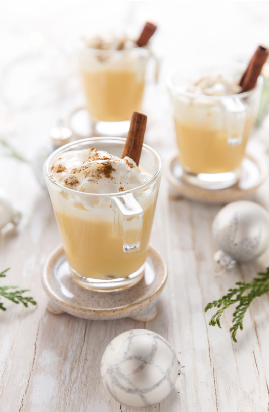 Glass mugs of eggnog with whipped cream on top and cinnamon sticks.