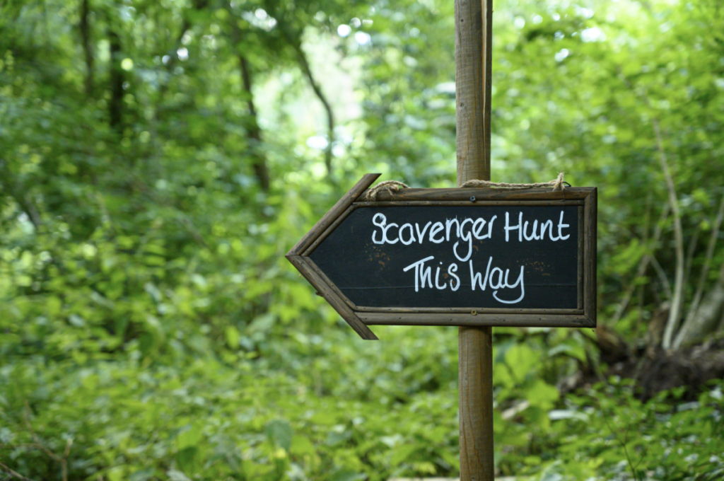 Arrow sign in the woods that says "Scavenger Hunt This Way."