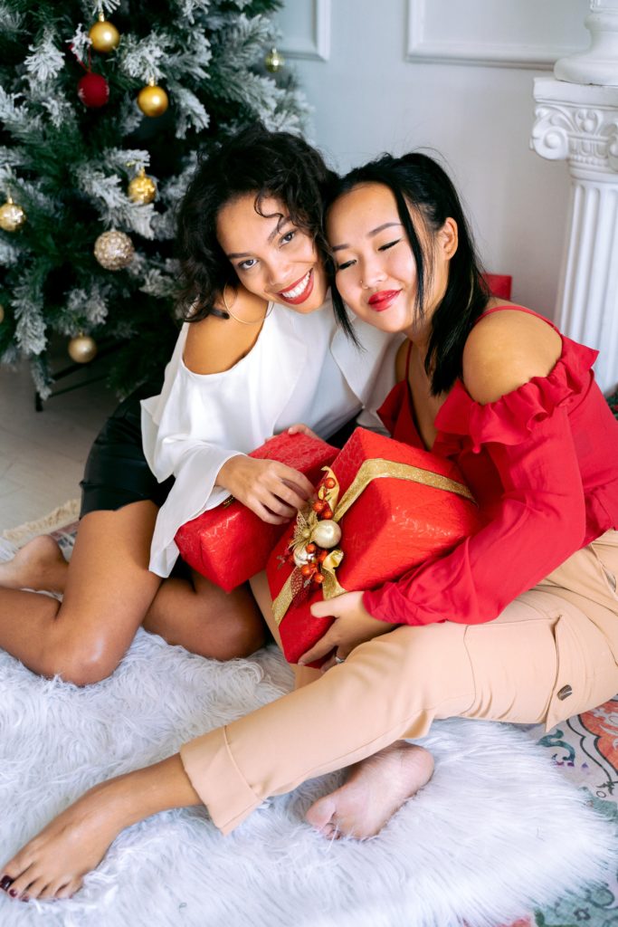 Two women sitting near a Christmas tree smiling and holding red, wrapped presents with gold ribbon.